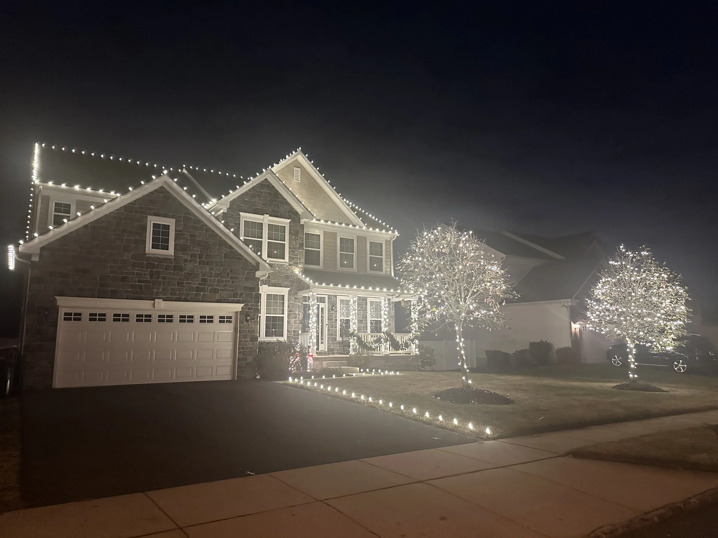 White Christmas lights along a two-story home’s roofline, front porch, walkway, and two front-yard trees